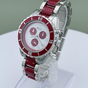 May include: A Geneva Platinum wristwatch with a silver-tone case and band, accented with a red bezel and red links. The watch face is white with silver numerals and three sub-dials. The watch is displayed on a clear stand.