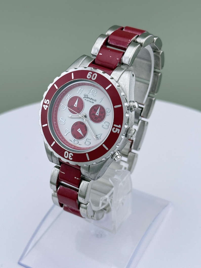 May include: A Geneva Platinum wristwatch with a silver-tone case and band, accented with a red bezel and red links. The watch face is white with silver numerals and three sub-dials. The watch is displayed on a clear stand.