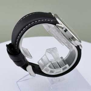 May include: A silver-toned wristwatch with a black leather band. The band is stitched with white thread. The watch is displayed on a clear plastic stand. The watch face is visible, and the watch is angled to show the side profile.