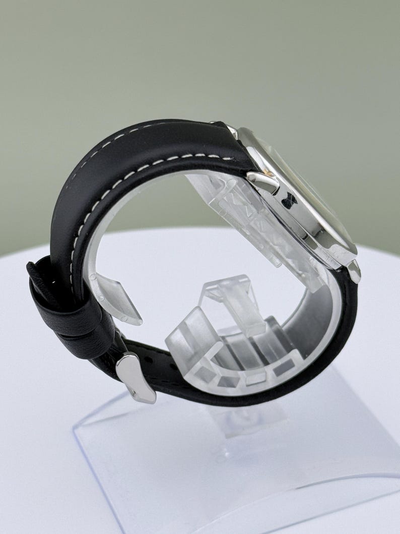 May include: A silver-toned wristwatch with a black leather band. The band is stitched with white thread. The watch is displayed on a clear plastic stand. The watch face is visible, and the watch is angled to show the side profile.
