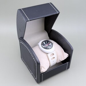 Puede incluir: Un reloj de pulsera blanco con esfera negra y roja, exhibido en una caja abierta de polipiel negra con costuras blancas. El reloj descansa sobre una almohada beige, mostrando su correa blanca y esfera redonda.