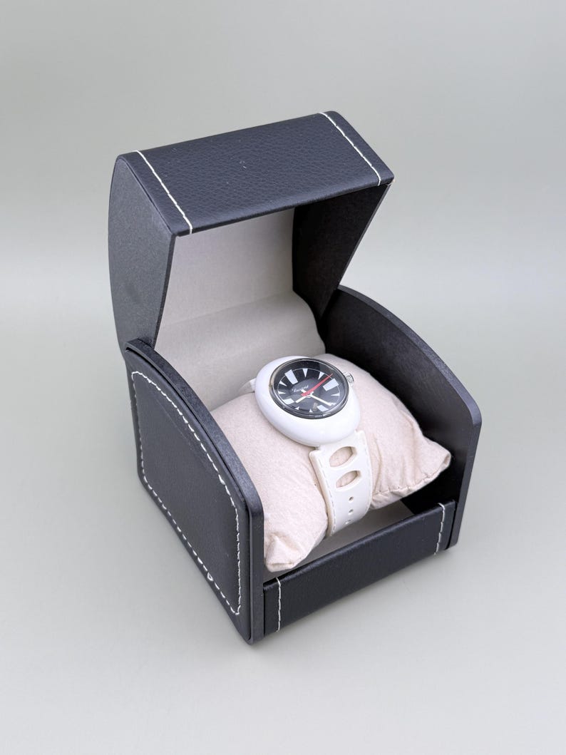 Puede incluir: Un reloj de pulsera blanco con esfera negra y roja, exhibido en una caja abierta de polipiel negra con costuras blancas. El reloj descansa sobre una almohada beige, mostrando su correa blanca y esfera redonda.