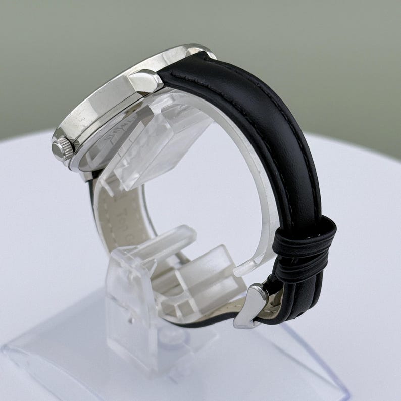 May include: A silver-toned wristwatch with a black leather band. The watch face is round and has a silver bezel. The black band is secured with a silver buckle. The watch is displayed on a clear plastic stand.