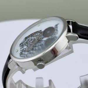 May include: Silver-toned wristwatch with a black leather band. The watch face features a black and white owl design. The watch has a round face with a silver bezel and a black leather strap.
