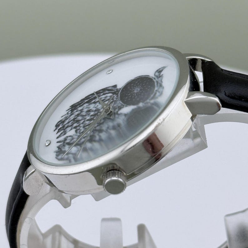 May include: Silver-toned wristwatch with a black leather band. The watch face features a black and white owl design. The watch has a round face with a silver bezel and a black leather strap.