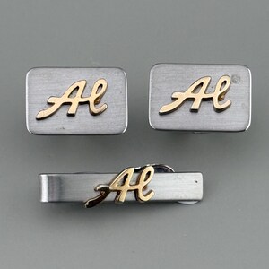 May include: A set of silver-tone cufflinks and a tie bar. Each piece features a brushed metal finish and a gold-colored cursive "A" and "H" monogram. The cufflinks are rectangular, and the tie bar is a slender, horizontal bar.