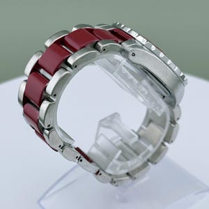 May include: A wristwatch with a stainless steel case and a burgundy-colored band. The band is composed of alternating stainless steel and burgundy links. The watch is displayed on a clear plastic stand, showcasing its design and materials.