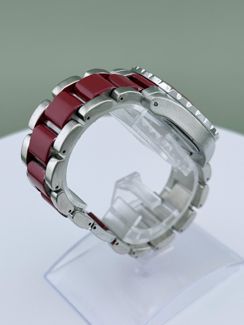 May include: A wristwatch with a stainless steel case and a burgundy-colored band. The band is composed of alternating stainless steel and burgundy links. The watch is displayed on a clear plastic stand, showcasing its design and materials.