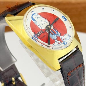 May include: Vintage gold-tone wristwatch with a brown leather band. The watch face features a cartoon character in a red and white circle with the text "99 Spiro Original". The watch has a rectangular gold case and a brown leather strap.