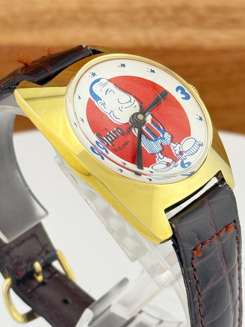 May include: Vintage gold-tone wristwatch with a brown leather band. The watch face features a cartoon character in a red and white circle with the text "99 Spiro Original". The watch has a rectangular gold case and a brown leather strap.
