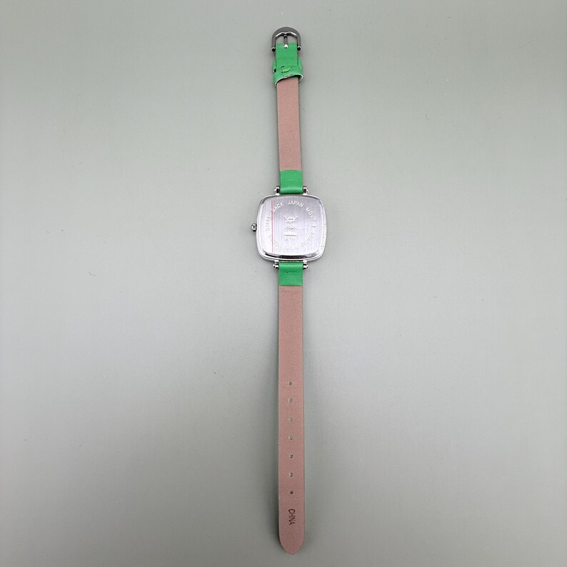 May include: A silver wristwatch with a square face and a light pink leather strap. The watch has green leather accents at the band attachments and a green buckle. The watch face has text that is difficult to read.