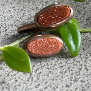 May include: A pair of oval cufflinks with a bronze-colored metal frame. The center of each cufflink has a sparkling, reddish-brown, glittery appearance. The cufflinks are resting on a gray surface with green leaves.