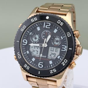K&ouml;nnte beinhalten: Eine goldfarbene Armbanduhr mit schwarzer L&uuml;nette und schwarzem Zifferblatt. Die Uhr verf&uuml;gt &uuml;ber digitale Anzeigen f&uuml;r Uhrzeit und Datum, mit dem Markennamen "Sauer" sichtbar. Das Armband der Uhr ist ebenfalls goldfarben.