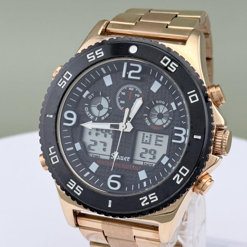 K&ouml;nnte beinhalten: Eine goldfarbene Armbanduhr mit schwarzer L&uuml;nette und schwarzem Zifferblatt. Die Uhr verf&uuml;gt &uuml;ber digitale Anzeigen f&uuml;r Uhrzeit und Datum, mit dem Markennamen "Sauer" sichtbar. Das Armband der Uhr ist ebenfalls goldfarben.