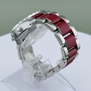 May include: A silver wristwatch with a burgundy band. The watch face is silver with black details. The band is composed of silver links and burgundy-colored rectangular segments. The watch is displayed on a clear plastic stand.