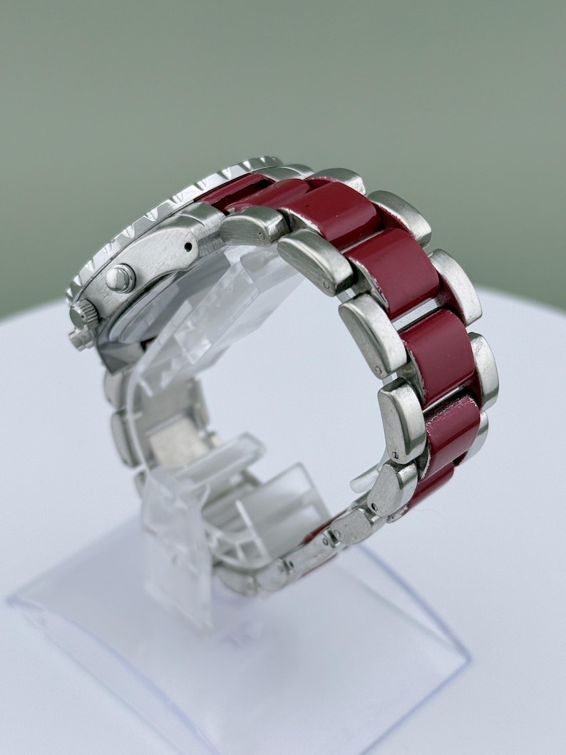 May include: A silver wristwatch with a burgundy band. The watch face is silver with black details. The band is composed of silver links and burgundy-colored rectangular segments. The watch is displayed on a clear plastic stand.