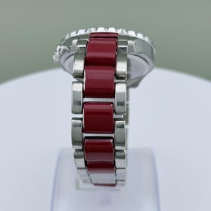 May include: A silver wristwatch with a red band. The watch face is silver with a black bezel. The band is composed of rectangular red links and silver links. The watch is displayed on a white surface.