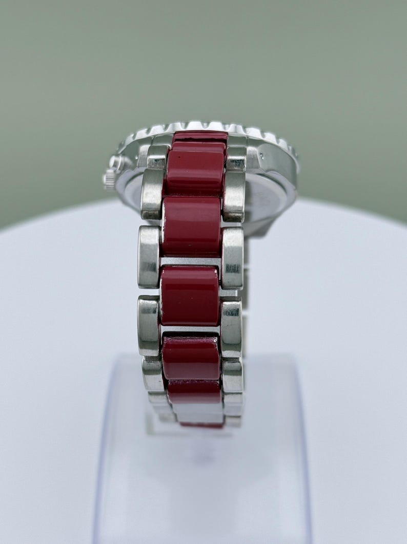 May include: A silver wristwatch with a red band. The watch face is silver with a black bezel. The band is composed of rectangular red links and silver links. The watch is displayed on a white surface.