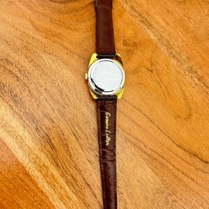 May include: A gold-toned vintage watch with a rectangular face and a brown leather band. The watch has a dark brown leather strap with a gold buckle. The back of the watch is silver-toned. The words "Genuine Leather" are printed on the strap.