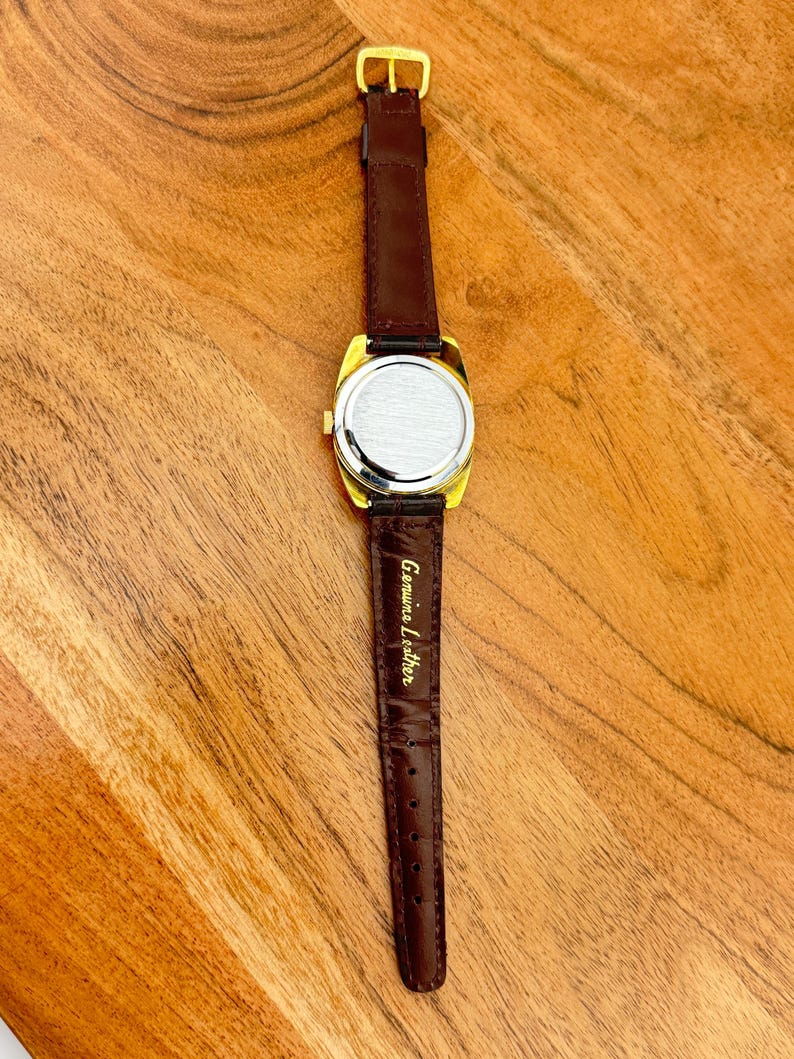 May include: A gold-toned vintage watch with a rectangular face and a brown leather band. The watch has a dark brown leather strap with a gold buckle. The back of the watch is silver-toned. The words "Genuine Leather" are printed on the strap.