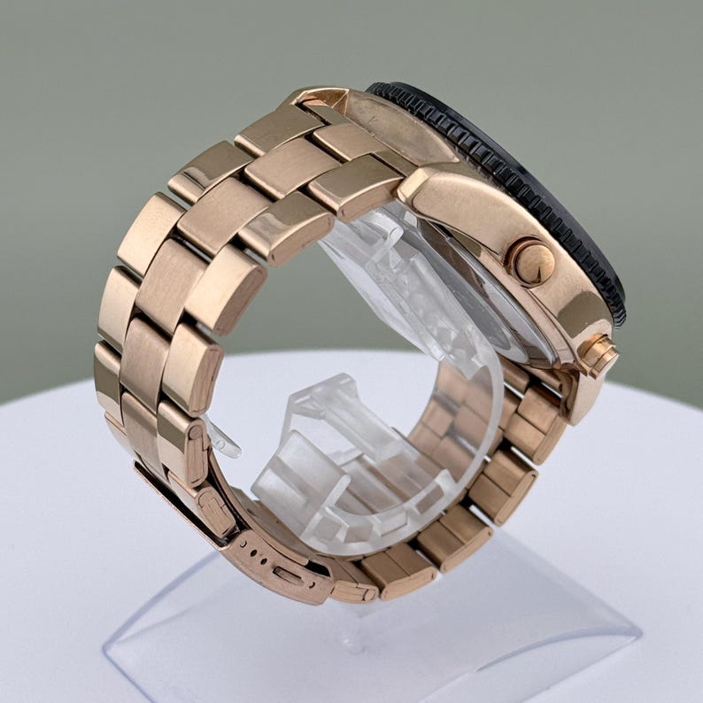 K&ouml;nnte beinhalten: Eine ros&eacute;goldfarbene Armbanduhr mit schwarzer L&uuml;nette und passendem Gliederarmband. Die Uhr wird schr&auml;g dargestellt, um die Details des Armbands und des Zifferblatts zu zeigen. Die Uhr steht auf einem transparenten St&auml;nder.