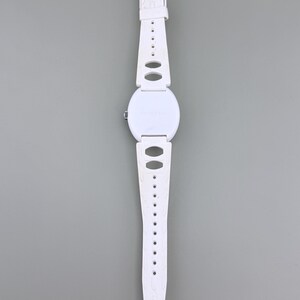 Puede incluir: Un reloj de pulsera blanco con esfera redonda y correa blanca a juego. La correa presenta cortes ovalados y un cierre de hebilla. El reloj se muestra sobre un fondo gris.