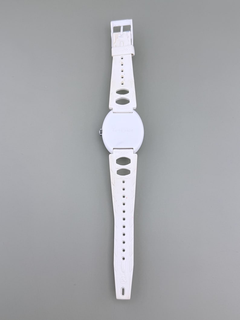Puede incluir: Un reloj de pulsera blanco con esfera redonda y correa blanca a juego. La correa presenta cortes ovalados y un cierre de hebilla. El reloj se muestra sobre un fondo gris.