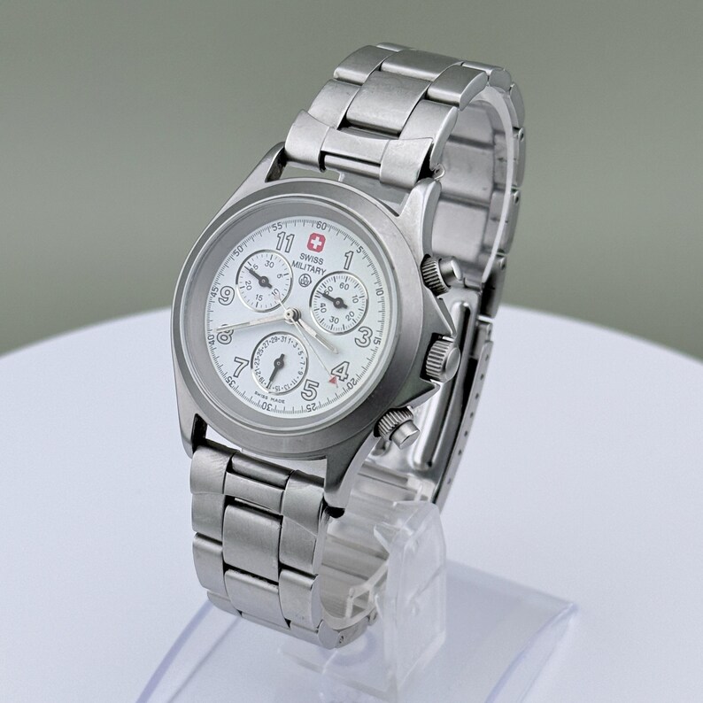 Puede incluir: Un reloj Swiss Military de acero inoxidable con esfera blanca y m&uacute;ltiples subesferas. El reloj presenta una correa plateada y un emblema de la bandera suiza. El reloj se exhibe sobre un soporte transparente.
