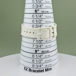 Puede incluir: Una correa de reloj color crema se muestra en una herramienta de medici&oacute;n blanca EZ Bracelet Mini. La herramienta mide de 12,7 cm a 22,2 cm. La correa del reloj tiene un cierre de hebilla.
