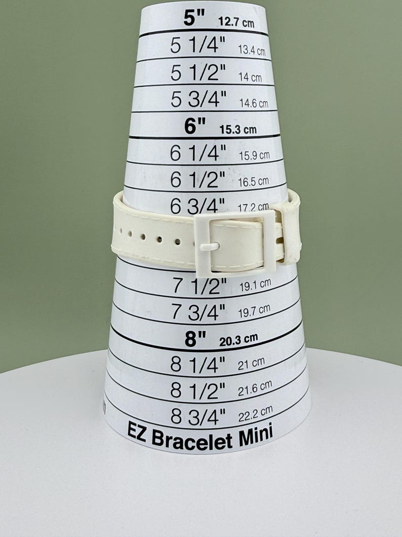 Puede incluir: Una correa de reloj color crema se muestra en una herramienta de medici&oacute;n blanca EZ Bracelet Mini. La herramienta mide de 12,7 cm a 22,2 cm. La correa del reloj tiene un cierre de hebilla.