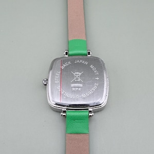 May include: A stainless steel watch with a square silver face and a green leather strap. The watch back is visible, with text that reads "STEEL BACK JAPAN MOVT SR626SW". The watch band is a light pink colour.