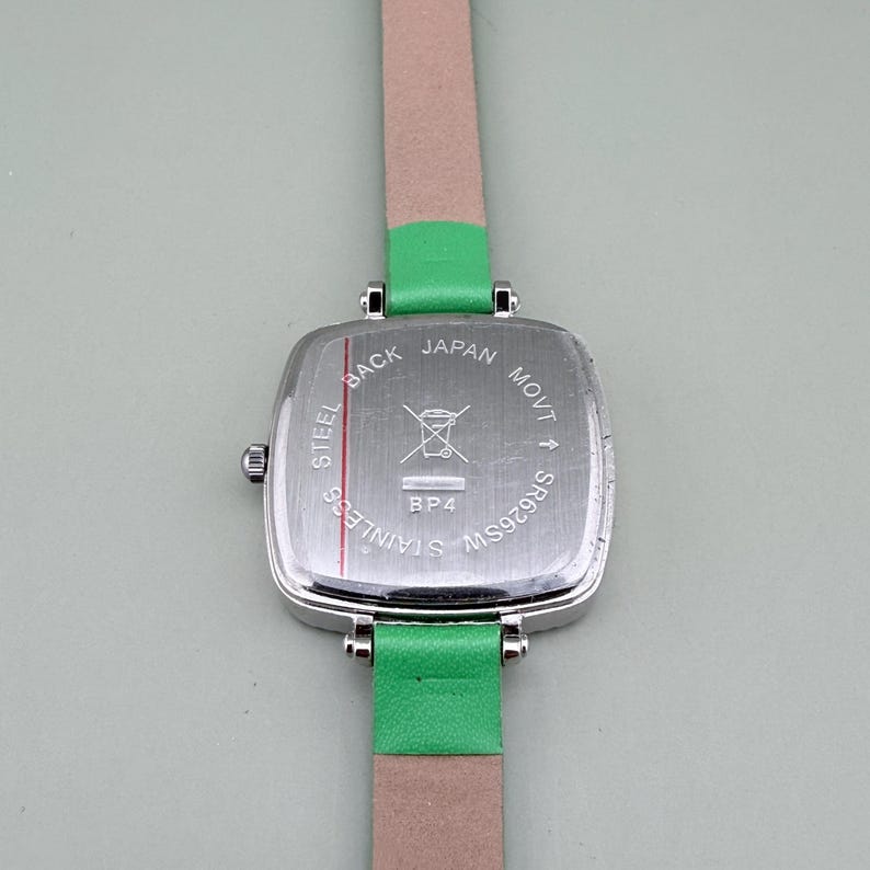 May include: A stainless steel watch with a square silver face and a green leather strap. The watch back is visible, with text that reads "STEEL BACK JAPAN MOVT SR626SW". The watch band is a light pink colour.