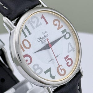 May include: A Swanie Quartz wristwatch with a white face, featuring multi-colored numerals and hands. The watch has a silver-toned case, a black leather band, and the text "JAPAN MOVT".