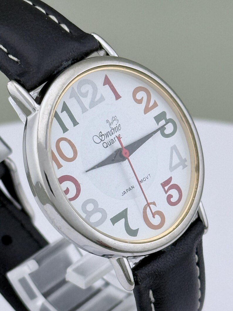 May include: A Swanie Quartz wristwatch with a white face, featuring multi-colored numerals and hands. The watch has a silver-toned case, a black leather band, and the text "JAPAN MOVT".