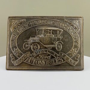 May include: A rectangular, antique-style metal belt buckle with an embossed design. The design features a vintage car within an oval, surrounded by text that reads "HENRY FORD DETROIT RECORD MODEL AUTOMOBILES YEAR."