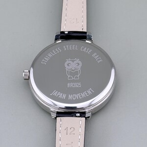 May include: Close-up of a silver wristwatch with a black leather band. The watch back is engraved with an owl illustration, the text "STAINLESS STEEL CASE BACK", "#IR3925", and "JAPAN MOVEMENT". The band is attached with a silver clasp.