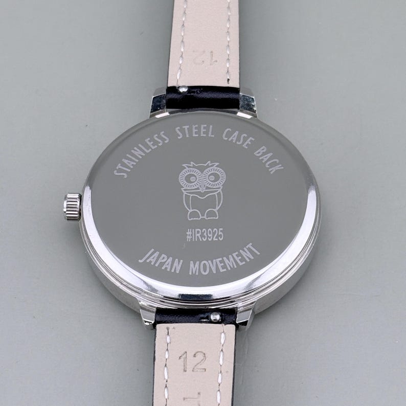 May include: Close-up of a silver wristwatch with a black leather band. The watch back is engraved with an owl illustration, the text "STAINLESS STEEL CASE BACK", "#IR3925", and "JAPAN MOVEMENT". The band is attached with a silver clasp.