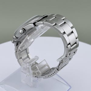 Puede incluir: Un reloj de pulsera plateado con una correa de acero inoxidable. El reloj se exhibe sobre un soporte de pl&aacute;stico transparente, mostrando el perfil lateral de la esfera y los eslabones de la correa. El reloj tiene una esfera redonda y un bisel texturizado.