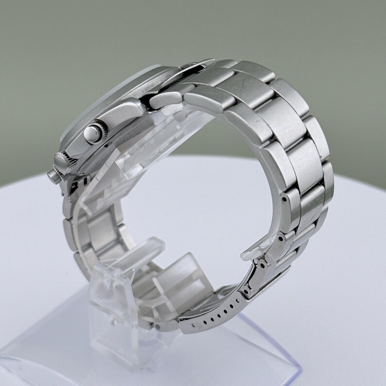 Puede incluir: Un reloj de pulsera plateado con una correa de acero inoxidable. El reloj se exhibe sobre un soporte de pl&aacute;stico transparente, mostrando el perfil lateral de la esfera y los eslabones de la correa. El reloj tiene una esfera redonda y un bisel texturizado.