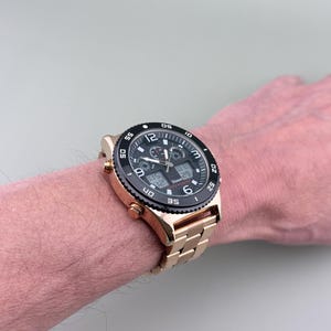 K&ouml;nnte beinhalten: Eine goldfarbene Armbanduhr mit schwarzer L&uuml;nette und schwarzem Zifferblatt. Die Uhr verf&uuml;gt &uuml;ber digitale Anzeigen und ein goldfarbenes Metallarmband. Die Uhr wird am Handgelenk getragen.