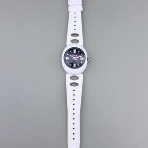 Puede incluir: Reloj de pulsera blanco con esfera en blanco y negro y correa blanca. La esfera del reloj tiene detalles en rojo y se ve el nombre de la marca. La correa tiene cortes ovalados y redondos.
