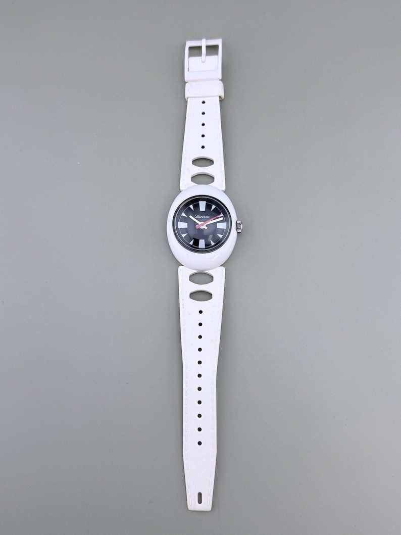 Puede incluir: Reloj de pulsera blanco con esfera en blanco y negro y correa blanca. La esfera del reloj tiene detalles en rojo y se ve el nombre de la marca. La correa tiene cortes ovalados y redondos.