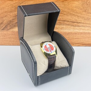 May include: A gold-toned wristwatch with a cartoon character on the face, set in a black leatherette box with white stitching. The watch has a brown leather band. The watch face is red and white.