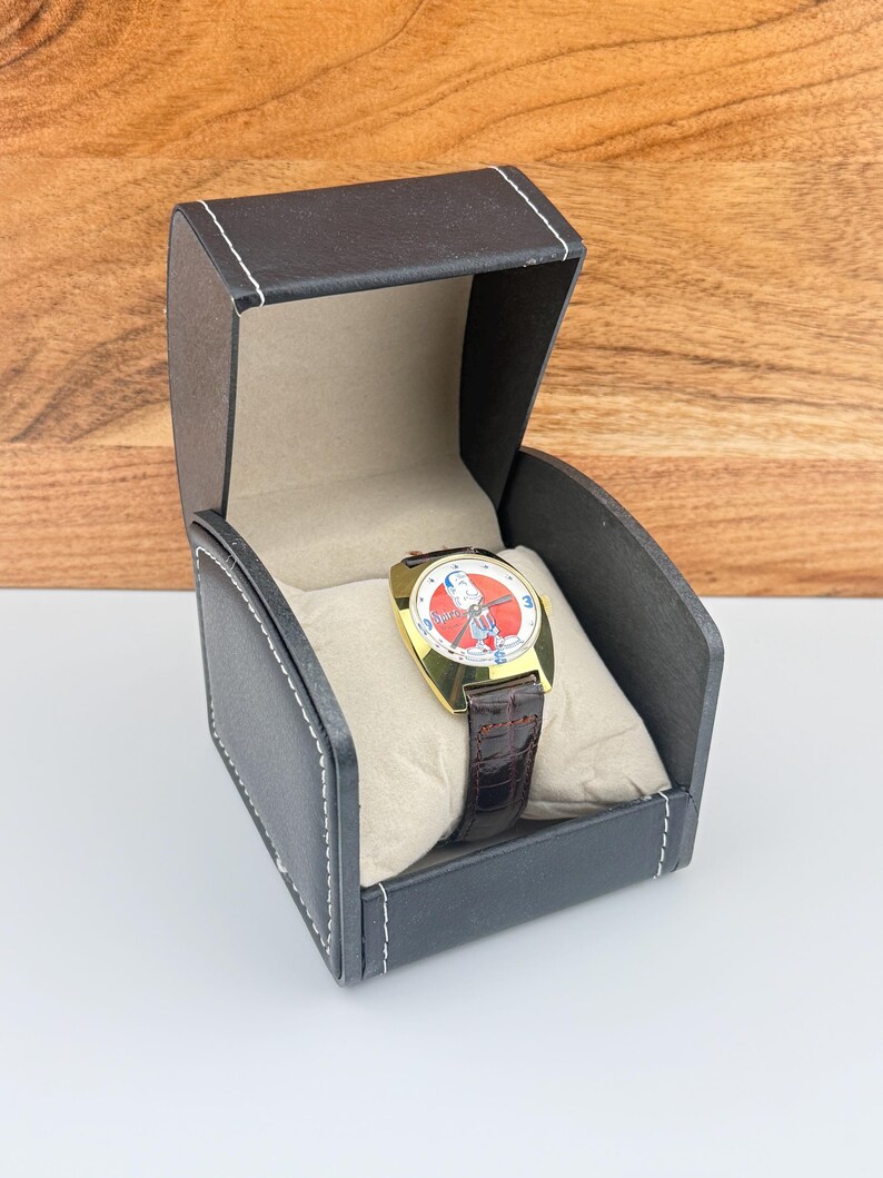 May include: A gold-toned wristwatch with a cartoon character on the face, set in a black leatherette box with white stitching. The watch has a brown leather band. The watch face is red and white.