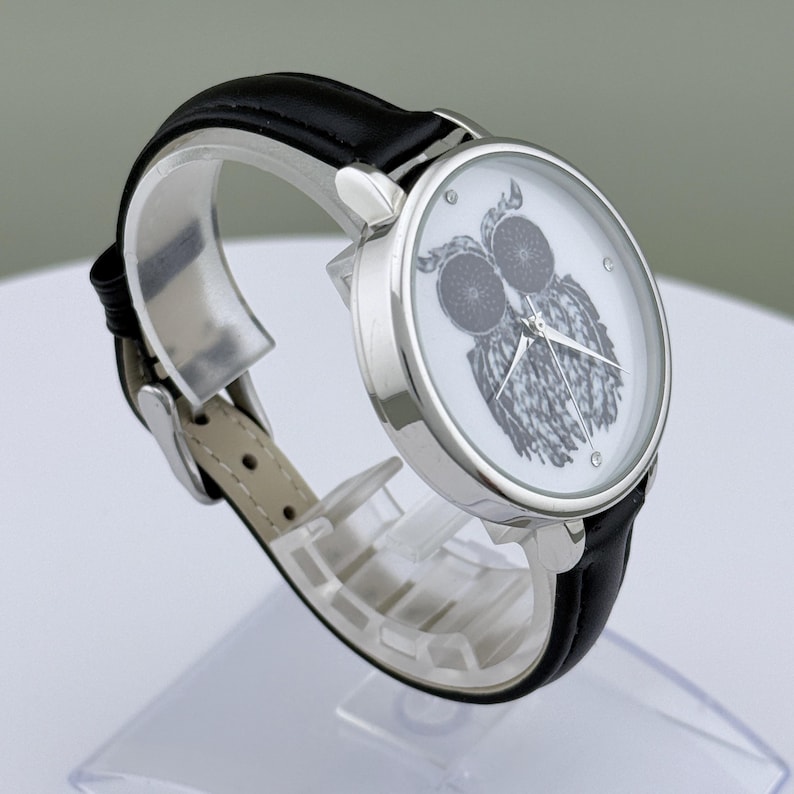 May include: A silver-toned wristwatch with a black leather band. The watch face features a detailed black and white owl illustration. The watch has a classic design with a buckle closure.