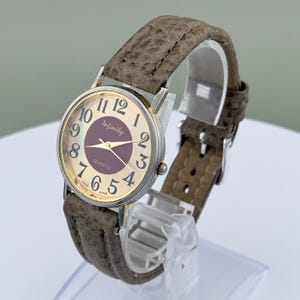 May include: A vintage wristwatch with a silver-tone case and a brown leather band. The watch face is gold with black numerals and a dark purple center. The watch reads "Infinity" and "Quartz".