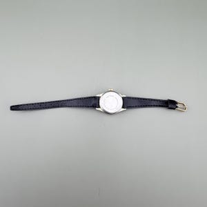 K&ouml;nnte beinhalten: Eine goldfarbene Uhr mit einem schwarzen Lederarmband. Das Zifferblatt der Uhr ist rund mit wei&szlig;em Hintergrund. Das Armband ist schmal und hat eine Schnalle. Die Uhr wird vor einem grauen Hintergrund gezeigt.