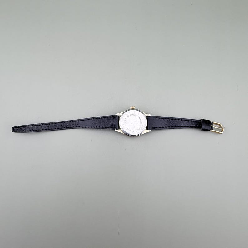 K&ouml;nnte beinhalten: Eine goldfarbene Uhr mit einem schwarzen Lederarmband. Das Zifferblatt der Uhr ist rund mit wei&szlig;em Hintergrund. Das Armband ist schmal und hat eine Schnalle. Die Uhr wird vor einem grauen Hintergrund gezeigt.