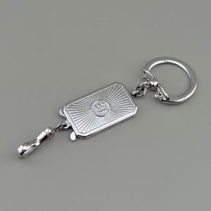 May include: Silver-toned keychain featuring a rectangular pendant with a sunburst design and a central emblem. The keychain includes a small handle and a circular key ring. The design is detailed and the metal has a polished finish.