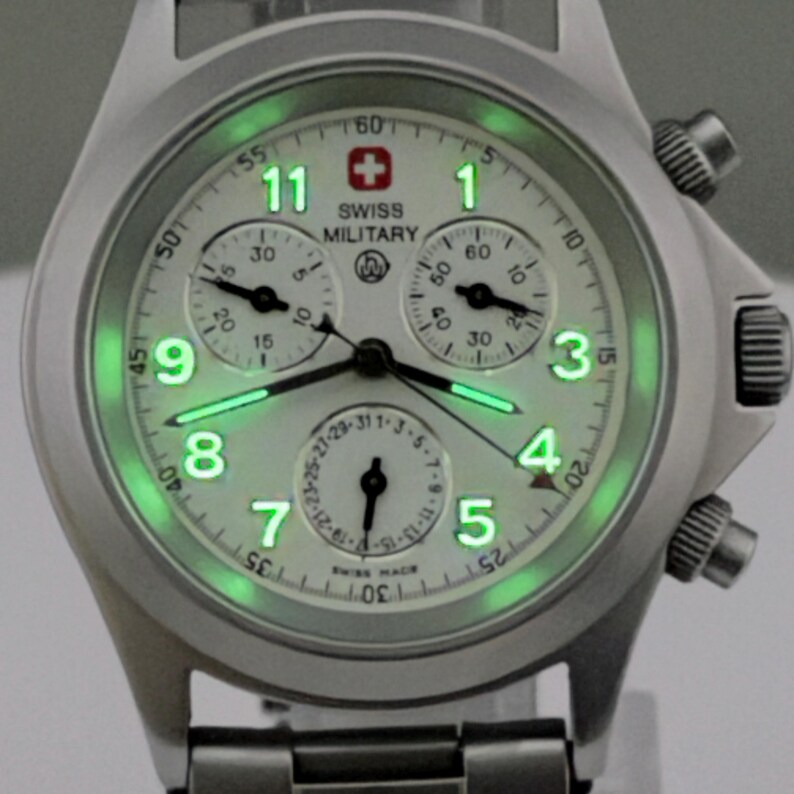 Puede incluir: Un reloj Swiss Military plateado con esfera blanca y manecillas y n&uacute;meros verdes luminosos. El reloj presenta tres subesferas, una pantalla de fecha y el logotipo Swiss Military. La correa es de acero inoxidable.